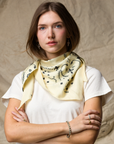 A woman with light brown hair crosses her arms and gazes forward, wearing a KUJTEN TIFA Scarf with black patterns over a white t-shirt, standing against a neutral, textured fabric backdrop.