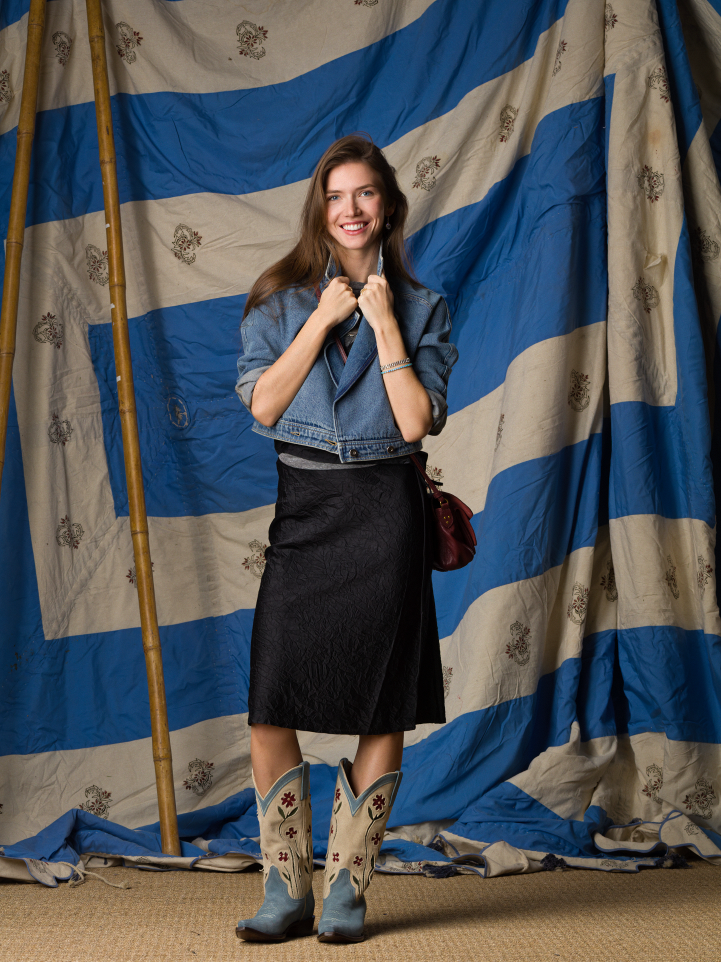 A smiling woman stands before a blue-and-white striped backdrop, wearing a Brazeau Tricot Crinkle Skirt, denim jacket, light blue cowboy boots, and holding her jacket collar with both hands.