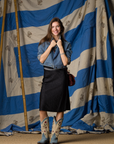 A smiling woman stands before a blue-and-white striped backdrop, wearing a Brazeau Tricot Crinkle Skirt, denim jacket, light blue cowboy boots, and holding her jacket collar with both hands.