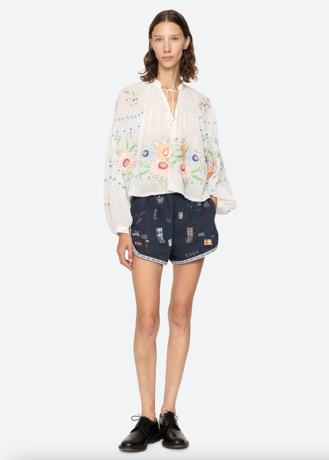 A woman stands facing forward in a sheer white floral blouse, Claira Shorts by Sea New York in a navy embroidered track-short style, and black oxford shoes.