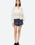 A woman stands facing forward in a sheer white floral blouse, Claira Shorts by Sea New York in a navy embroidered track-short style, and black oxford shoes.