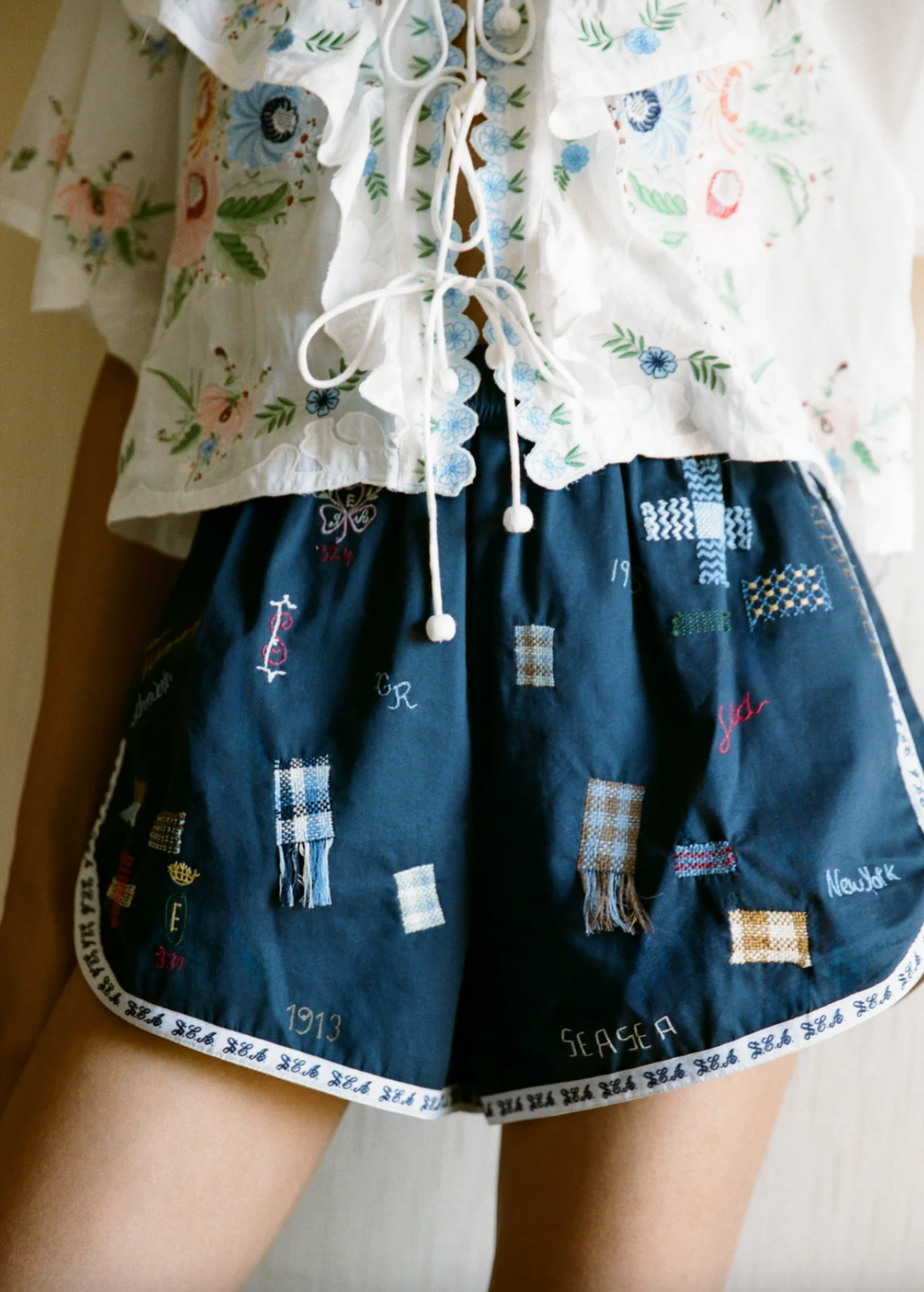 A person wears Sea New York’s Claira Shorts—navy blue, embroidered with colorful patches and an elastic waistband—paired with a white floral ruffled blouse. The image is cropped from mid-torso to mid-thigh.