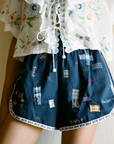 A person wears Sea New York’s Claira Shorts—navy blue, embroidered with colorful patches and an elastic waistband—paired with a white floral ruffled blouse. The image is cropped from mid-torso to mid-thigh.