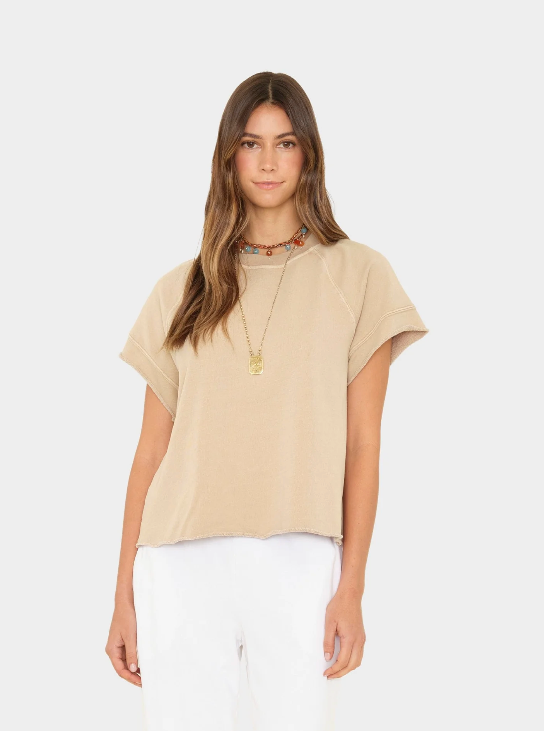 A woman with long brown hair wears the Xirena Callie Sweatshirt—a vintage-washed, boxy French terry style—paired with white pants and layered necklaces. She stands against a plain light background, gazing at the camera neutrally.