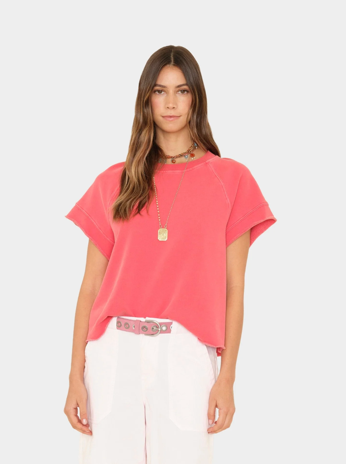 A woman with long brown hair wears the Xirena Callie Sweatshirt, a vintage washed French terry boxy fit in pink, paired with white pants and layered necklaces. She stands against a plain light gray background, looking at the camera.