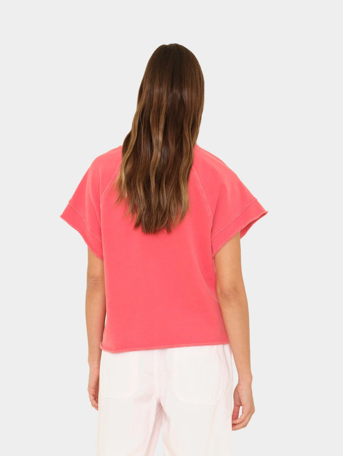 A woman with long brown hair, seen from the back, wears the Xirena Callie Sweatshirt in pink and white pants against a plain light background.