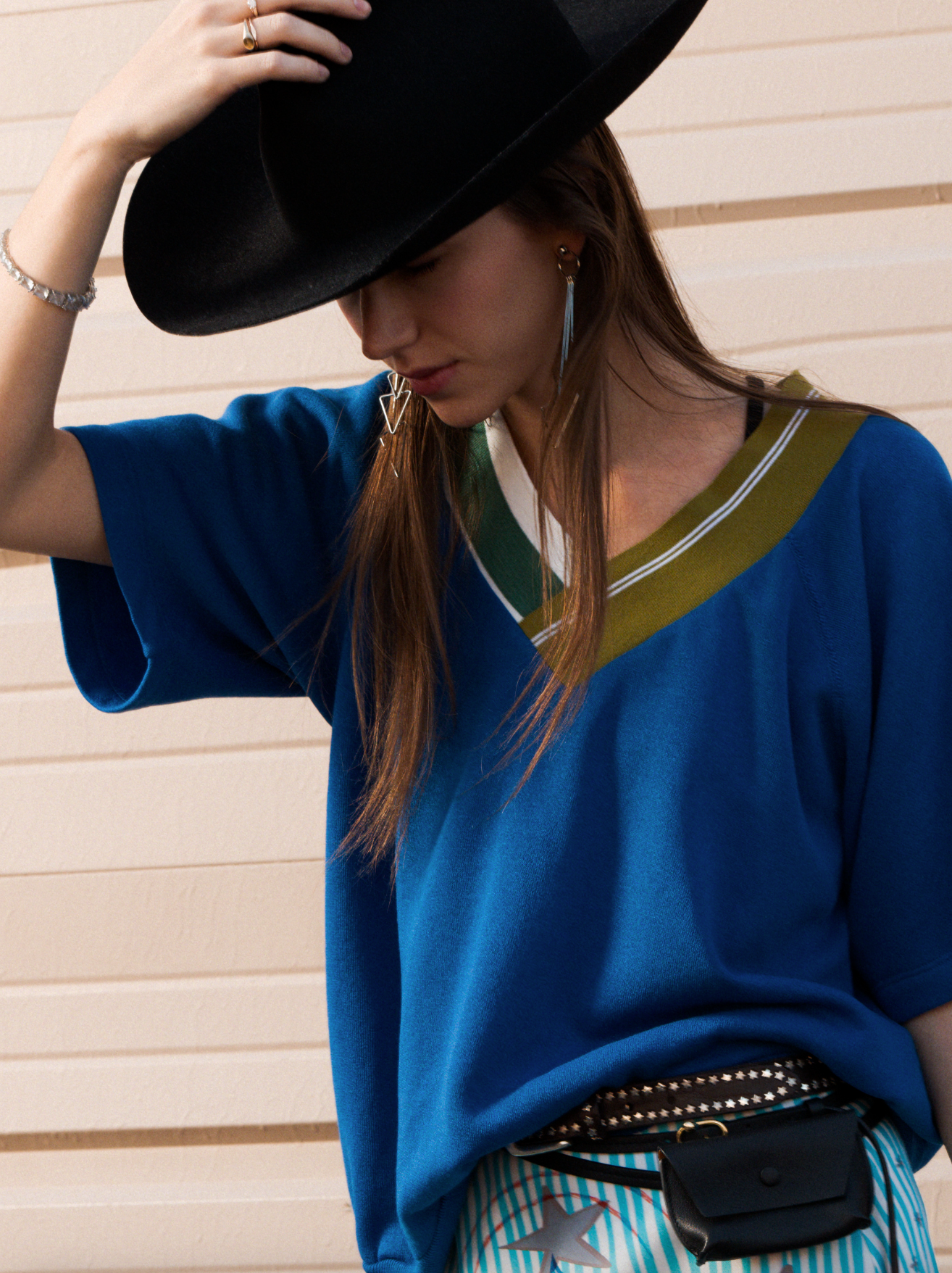 A woman in the SIDE CAR mixed rib vee top by Aquarius Cocktail, striped pants, and a wide-brimmed black hat stands against a beige wall, head tilted down so the hat partially shades her face. She adds earrings, rings, and a studded belt bag.