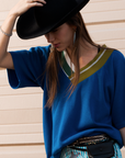 A woman in the SIDE CAR mixed rib vee top by Aquarius Cocktail, striped pants, and a wide-brimmed black hat stands against a beige wall, head tilted down so the hat partially shades her face. She adds earrings, rings, and a studded belt bag.