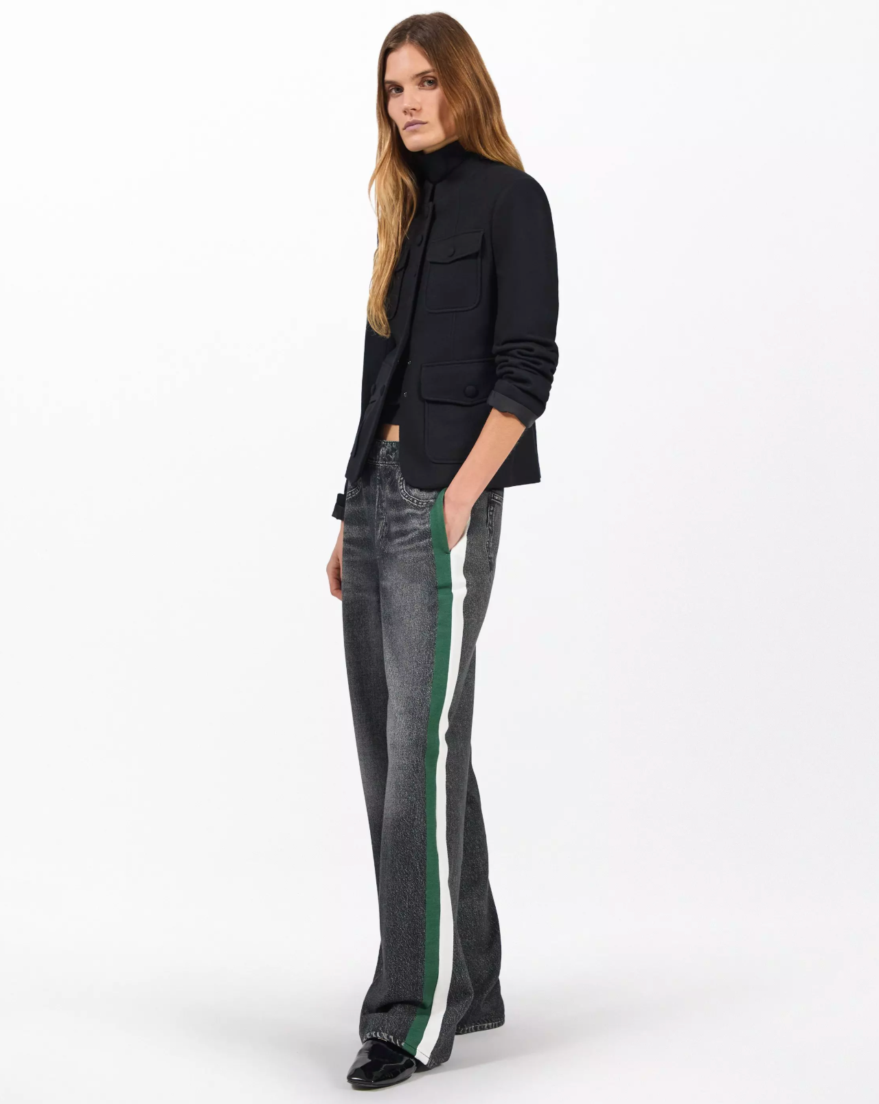 A person with long, light brown hair wears a black blazer, black top, and Rag & Bone's miramar terry track pant pull on with bold white and green side stripes, standing against a plain white background.