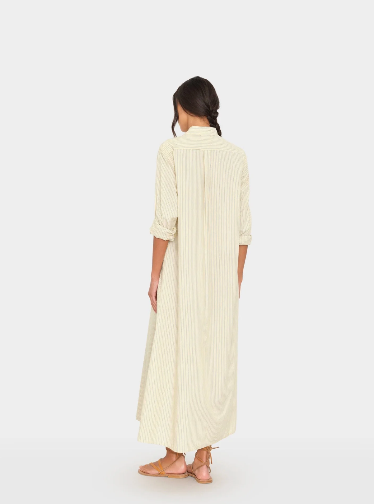 A woman with braided hair stands facing away, wearing the Xirena Boden Dress—a long, light cotton shirtdress with rolled sleeves and sandals—against a plain white background.