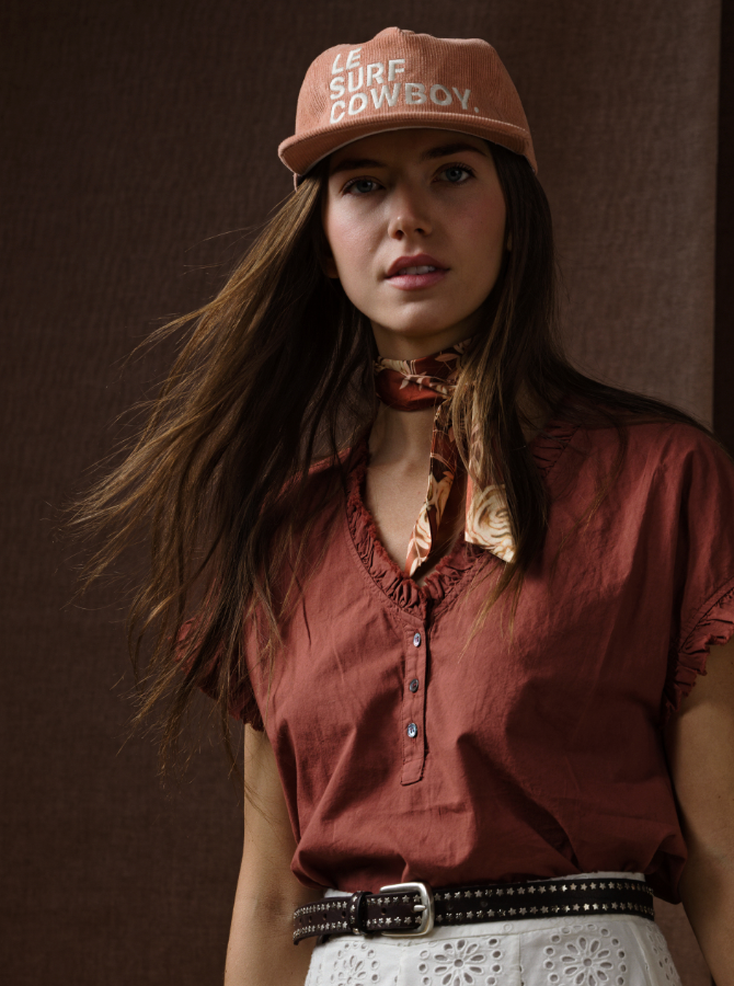 A woman stands in front of a brown textured background wearing the Xirena Darcy Top, a rust-colored cotton poplin blouse, paired with a pale pink "LE SURF COWBOY" cap, patterned neck scarf, and white eyelet skirt with a black belt.