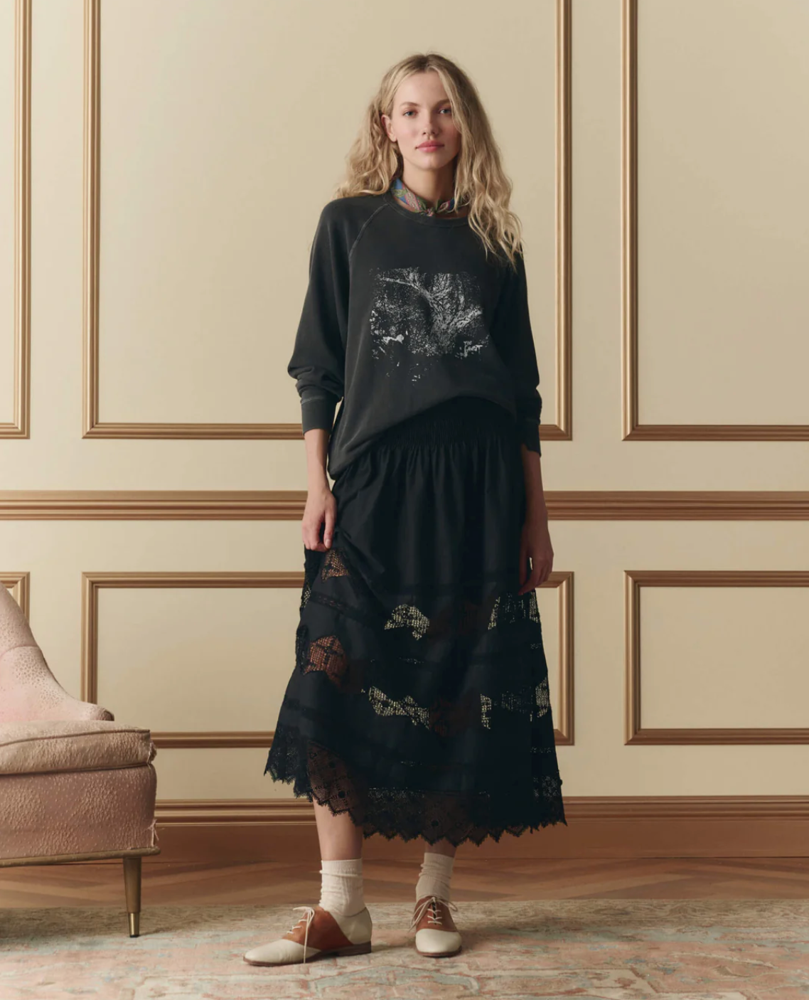 A woman stands indoors by a pink armchair and beige paneled wall, wearing The Rosette Crochet Canary Skirt by The Great Inc. with a black animal graphic sweatshirt, white socks, and brown-and-white oxford shoes.