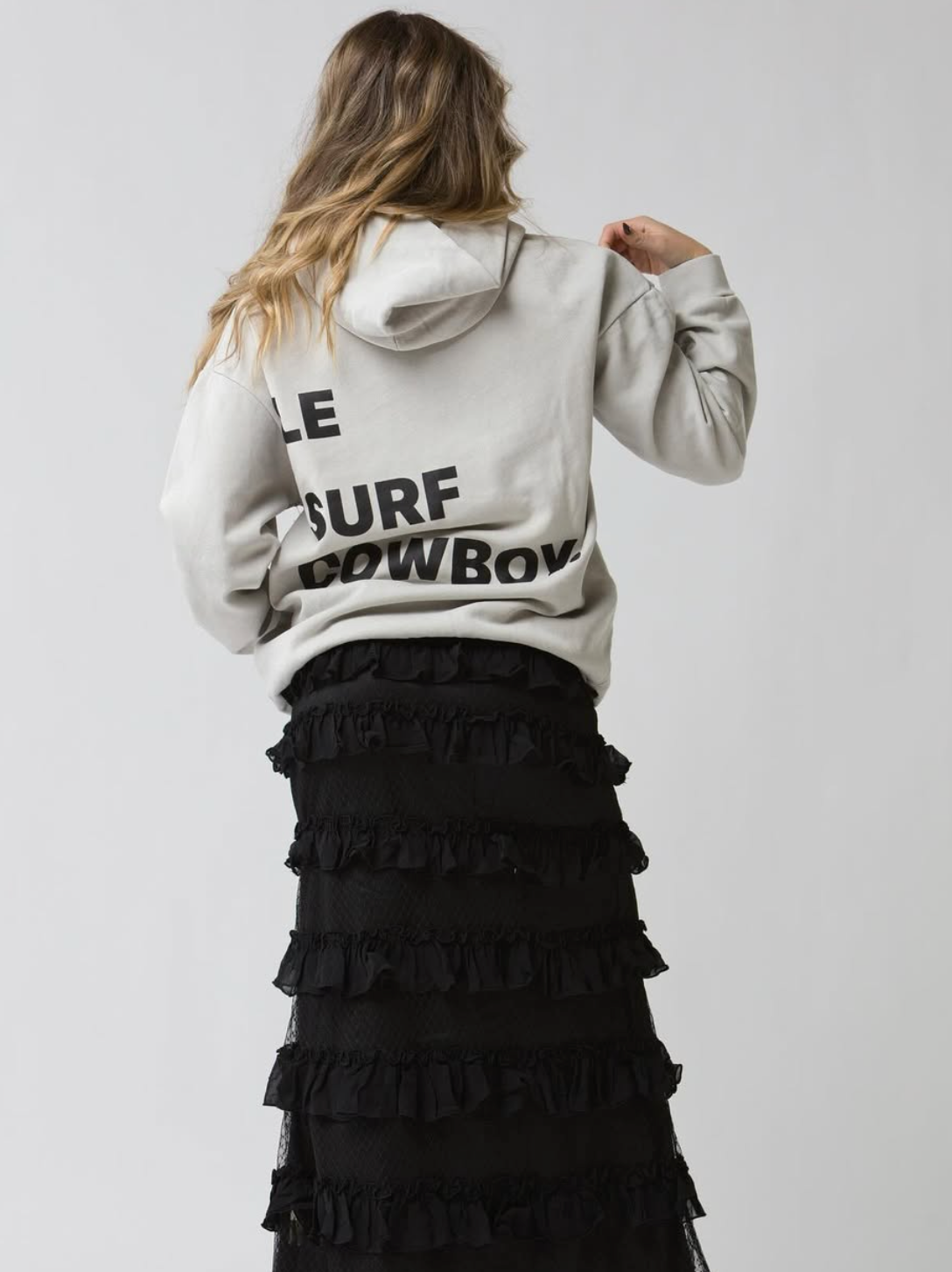 A person faces away in a light Ping Pong Surf Club Le Surf Cowboy Hoodie with bold black "LE SURF COWBOY" text, paired with a long black tiered ruffle skirt, set against a plain light background.