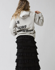 A person faces away in a light Ping Pong Surf Club Le Surf Cowboy Hoodie with bold black "LE SURF COWBOY" text, paired with a long black tiered ruffle skirt, set against a plain light background.