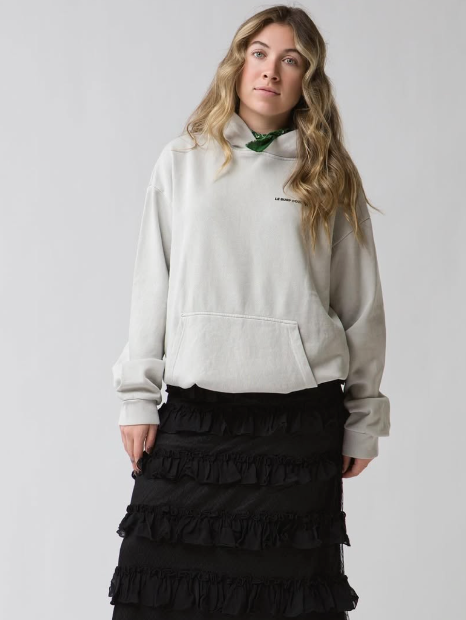 A woman with wavy blonde hair wears the Ping Pong Surf Club Le Surf Cowboy Hoodie in light gray, paired with a green scarf and a ruffled black skirt, standing against a plain light background.