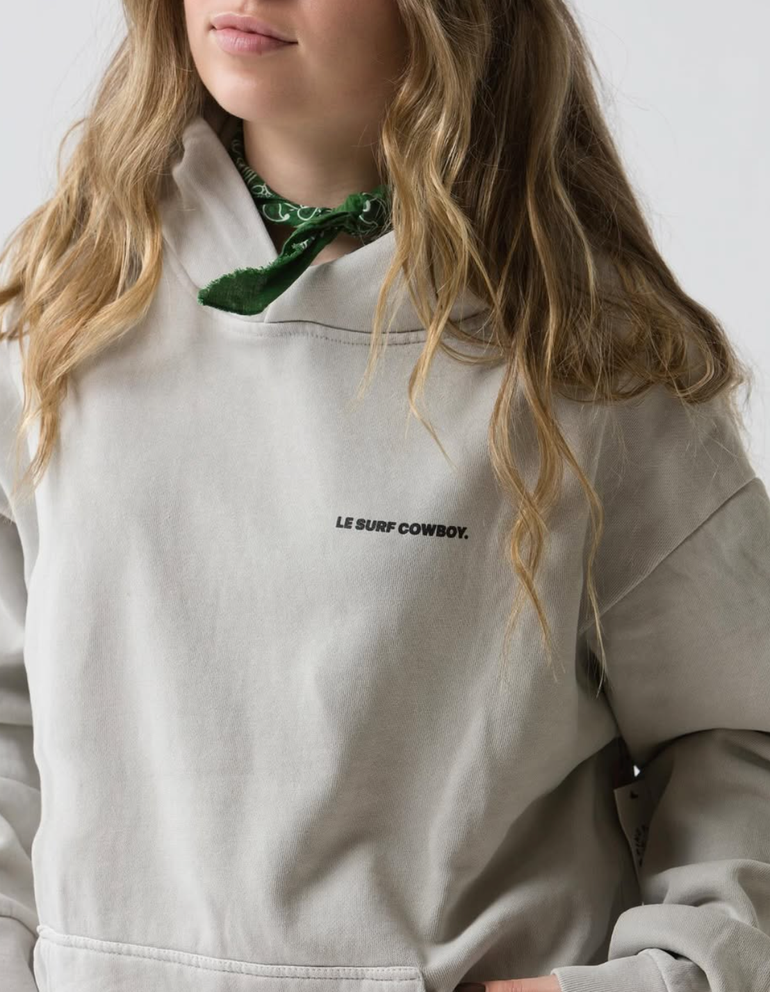 A person with long wavy hair wears the light-colored Le Surf Cowboy Hoodie by Ping Pong Surf Club, featuring "LE SURF COWBOY." on the chest and a green bandana. Their face is partially visible against a plain white background.