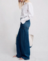 A person with long blonde hair, in a white button-up shirt and Denimist DECK PANT SHANE INDIGO wide-leg jeans, stands in profile against a plain light background, holding a large light tote bag and wearing sandals.