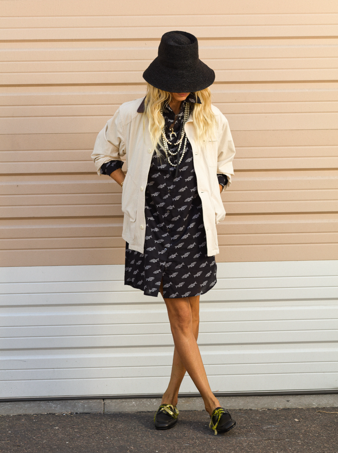 A person stands against a beige and white striped wall, hands in pockets, wearing CLOSED's patterned black Shirtdress paired with a wide-brimmed black hat, cream oversized jacket, layered necklaces, and black shoes with green details.