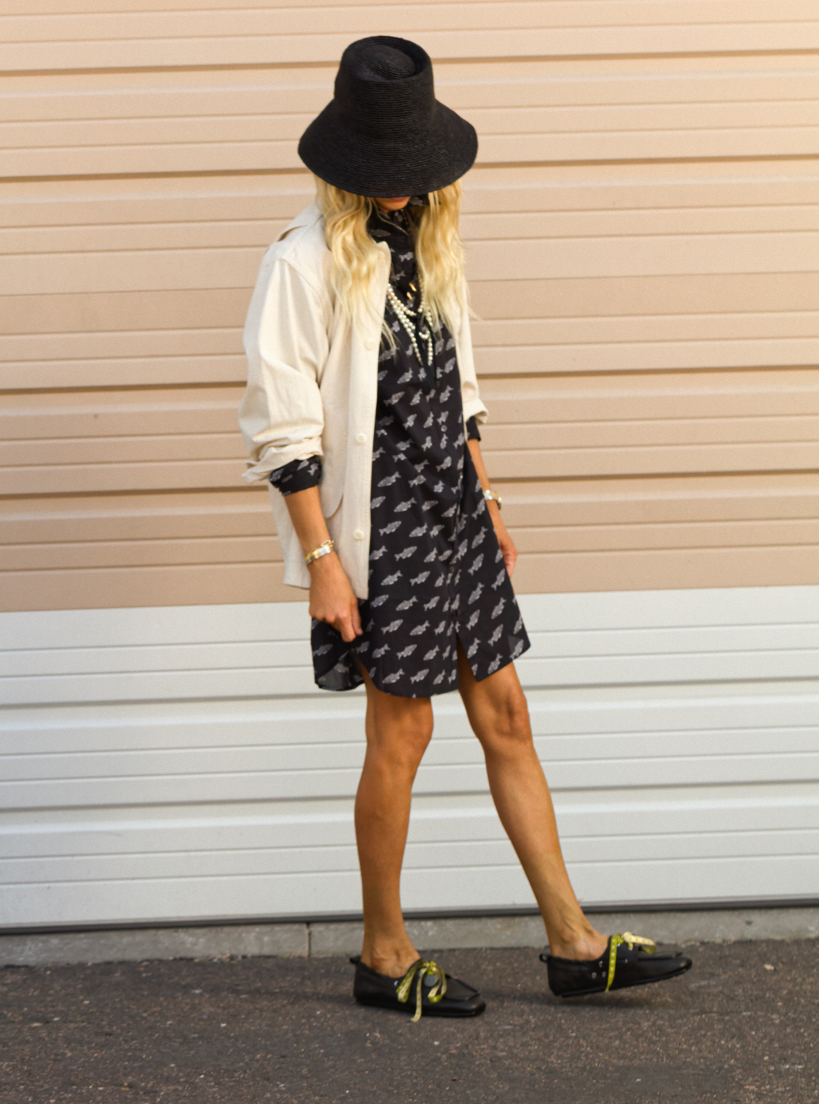 Wearing a CLOSED Shirtdress, black wide-brim hat, and black shoes with green laces, a person stands against a beige and white paneled wall. The hat covers their face.