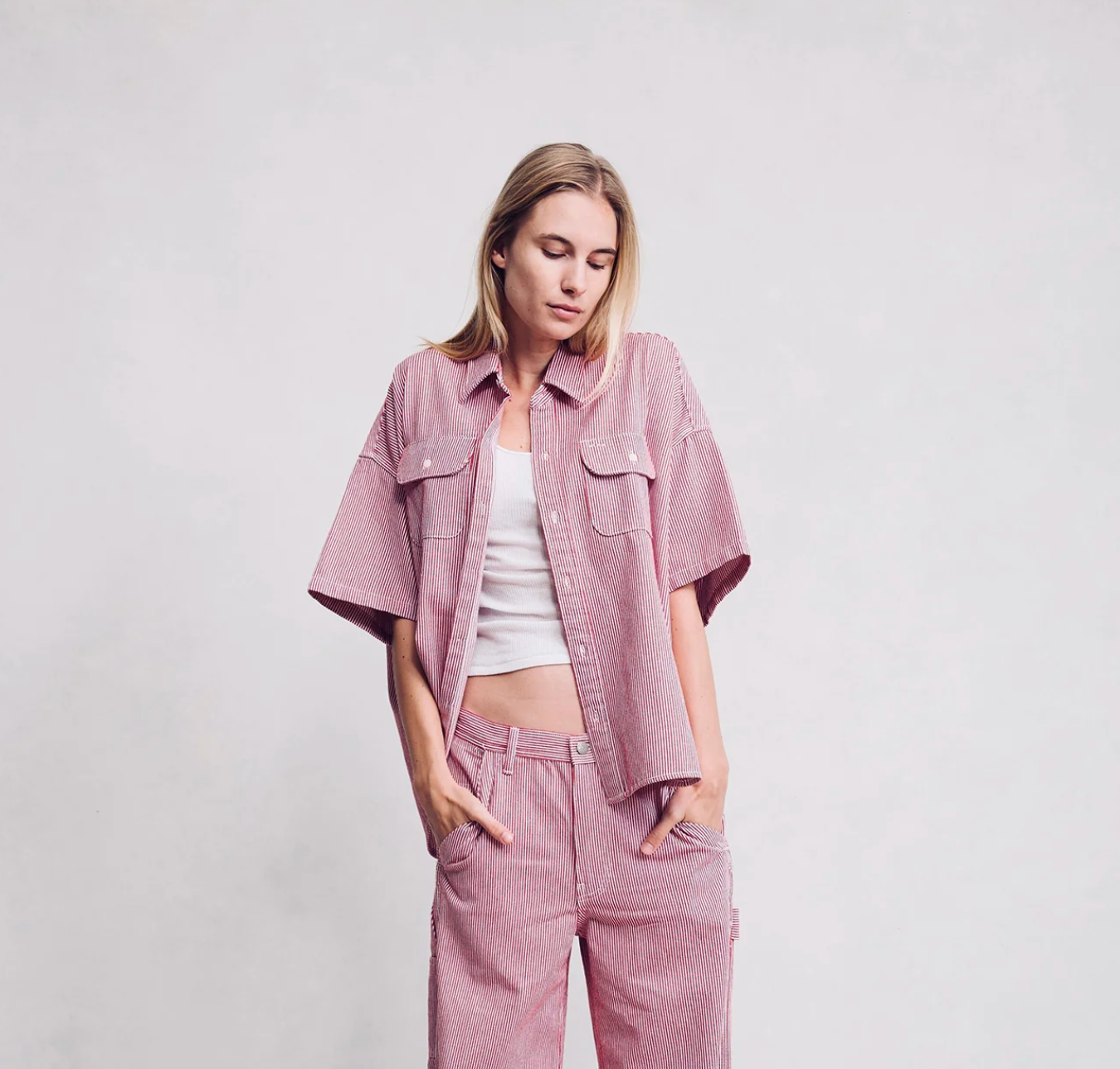A woman stands against a plain light background, wearing the Denimist Short Sleeve Button Down Workshirt with flap chest pockets over a white tank and matching pink pants. She looks down, hands in her pockets.