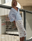 Cissa Summer Pencil Skirt in Venetian Embroidery with blue striped shirt, styled outdoors by a metal fence.