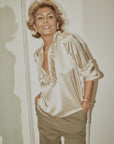 A woman with short, wavy blonde hair smiles with her hands in her khaki pockets, wearing the Celine Silk shirt by A Shirt Thing and gold jewelry as she stands indoors near a mirror.