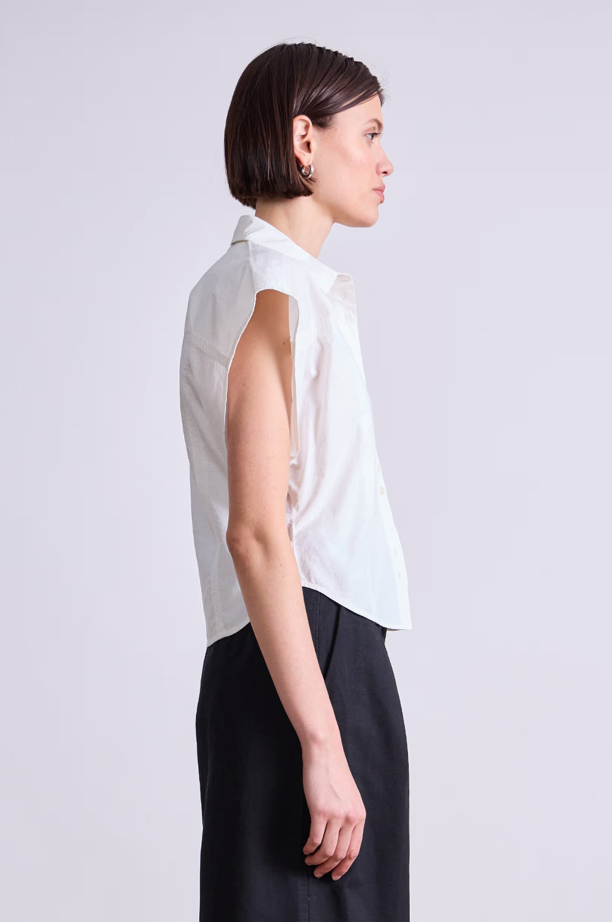 A person with short dark hair stands in profile, wearing the Apiece Apart Toni Sleeveless Top—an organic cotton white button-up shirt—paired with black pants, highlighting a modern look against a plain light background.