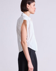 A person with short dark hair stands in profile, wearing the Apiece Apart Toni Sleeveless Top—an organic cotton white button-up shirt—paired with black pants, highlighting a modern look against a plain light background.
