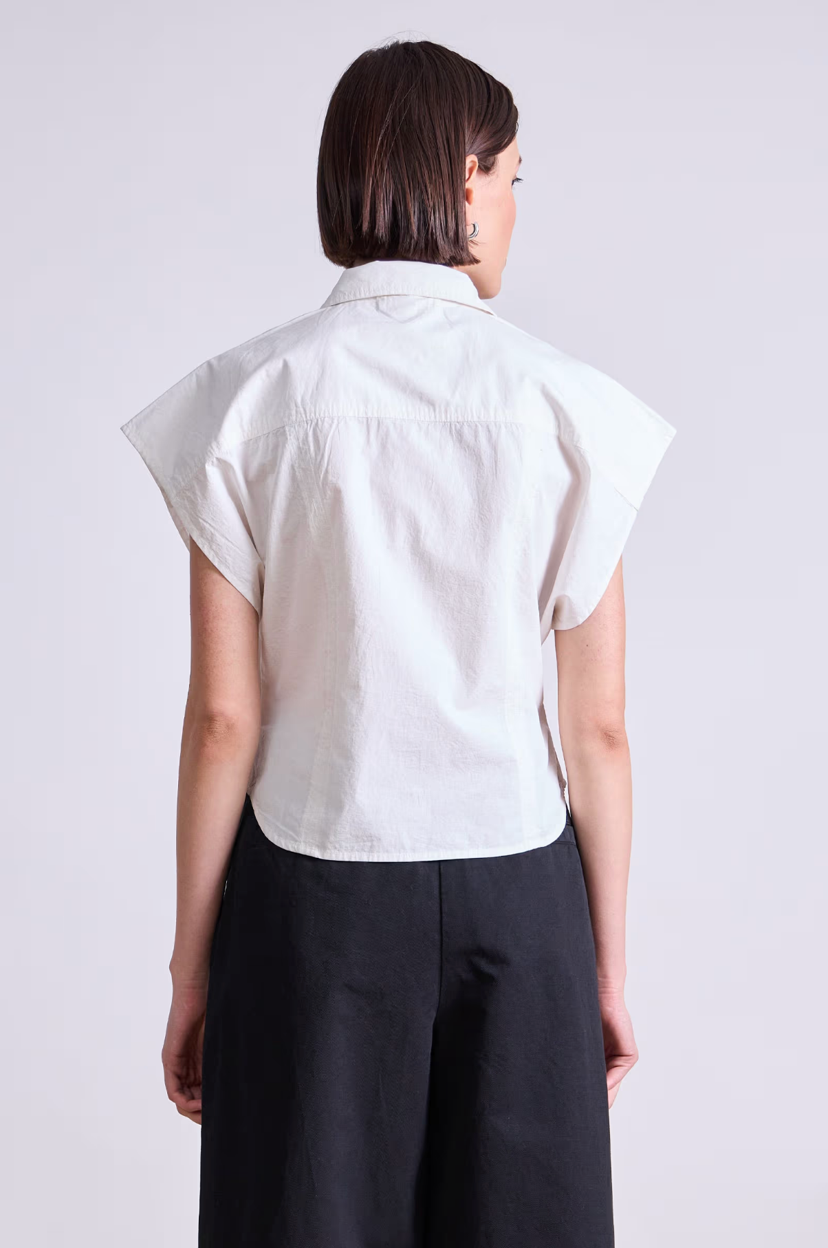 Seen from behind, a person with short brown hair wears the Apiece Apart Toni Sleeveless Top in white organic cotton and black high-waisted pants, creating a modern silhouette against a plain light background.