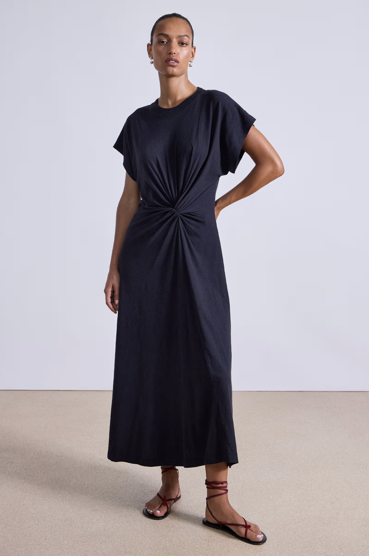 A woman stands against a plain background wearing the Apiece Apart Volta Twist Dress—an ankle-length, short-sleeved black organic cotton dress with a gathered twist waist—paired with brown strappy flat sandals.