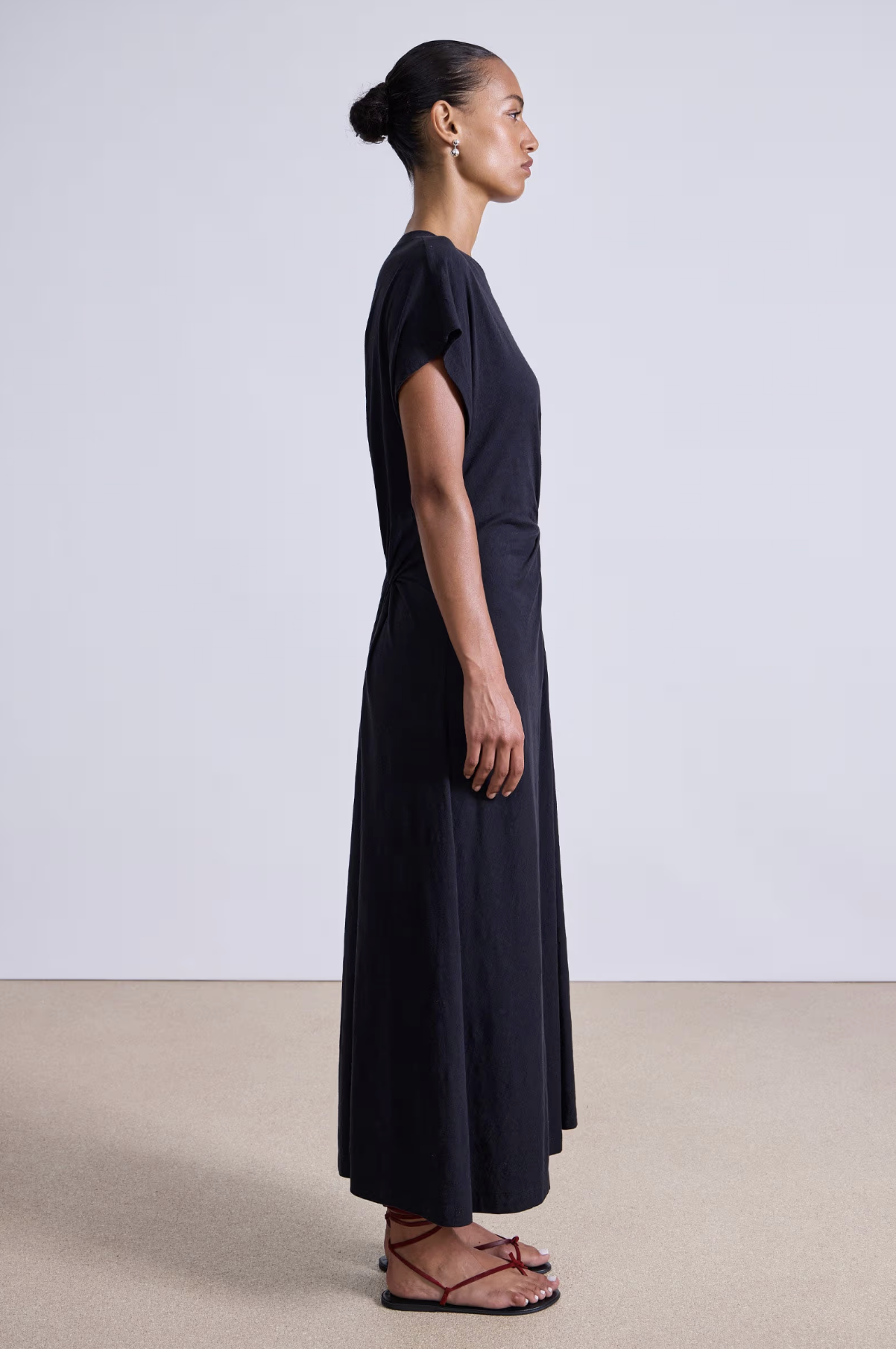 A woman in profile stands against a plain background, wearing the Apiece Apart Volta Twist Dress—an ankle-length, black organic cotton dress with short sleeves—and brown flat sandals. Her hair is styled in a neat bun.