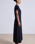 A woman in profile stands against a plain background, wearing the Apiece Apart Volta Twist Dress—an ankle-length, black organic cotton dress with short sleeves—and brown flat sandals. Her hair is styled in a neat bun.