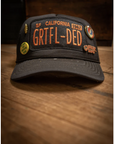 The MadeWorn Hat by Made Worn, a black baseball cap with "GRTFL–DED SF CALIFORNIA 1967" on the front, features limited edition pins and an adjustable back strap, displayed on a wooden surface.
