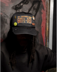 A person with long hair sits in shadow, wearing a Made Worn MadeWorn Hat with a custom front patch and limited edition pins reading "SF CALIFORNIA 1967 GRTFL-DED." The background is dark and abstract.