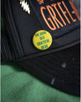 A close-up of the MadeWorn Hat by Made Worn, a black mesh cap with a custom "GRATEFUL DEAD" patch in orange and a limited edition "THE GOOD OLD GRATEFUL DEAD" pin, resting on green fabric.
