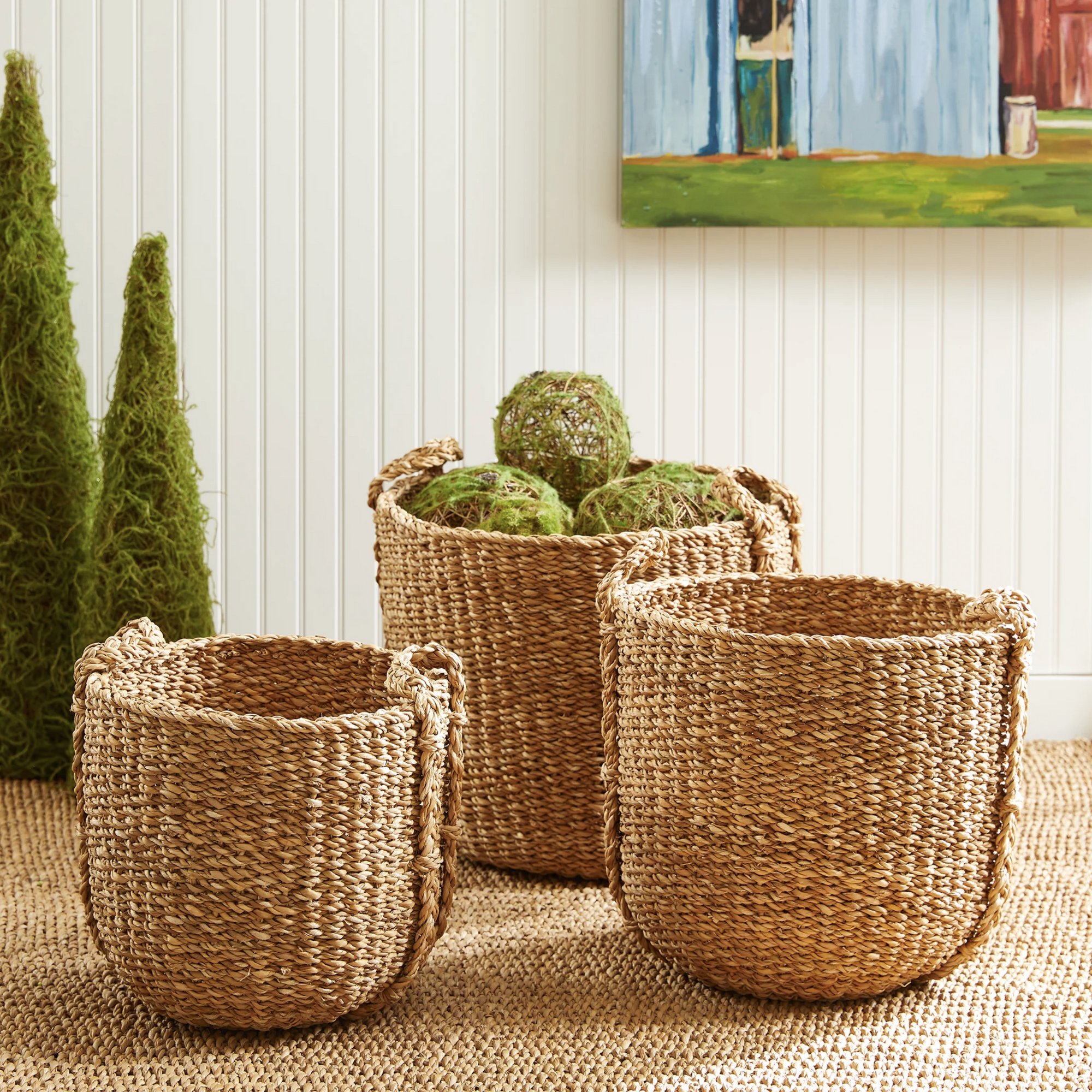 Three Napa Home & Garden Seagrass Round Drum Baskets rest on a textured rug, with the largest holding green moss balls. In the background, cone-shaped green topiaries and a colorful painting create a stylish storage display.