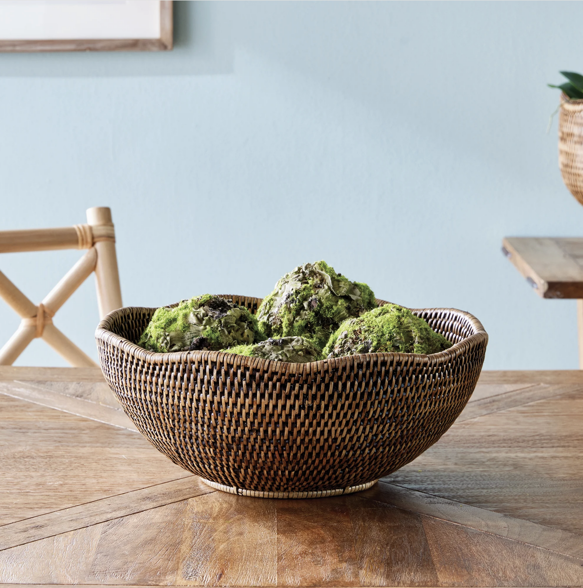 The Burma Rattan Scalloped Bowl by Napa Home & Garden, filled with decorative green moss rocks, rests on a wooden table with a chair and another wicker piece in the background.