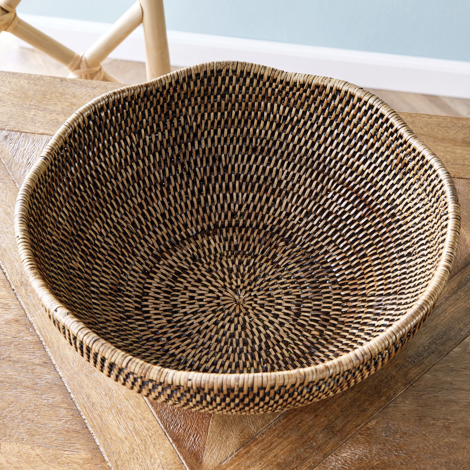 The Burma Rattan Scalloped Bowl by Napa Home & Garden, featuring a checkered brown and black woven design that reflects Burmese craftsmanship, sits empty on a wooden table with a light chair in the background.