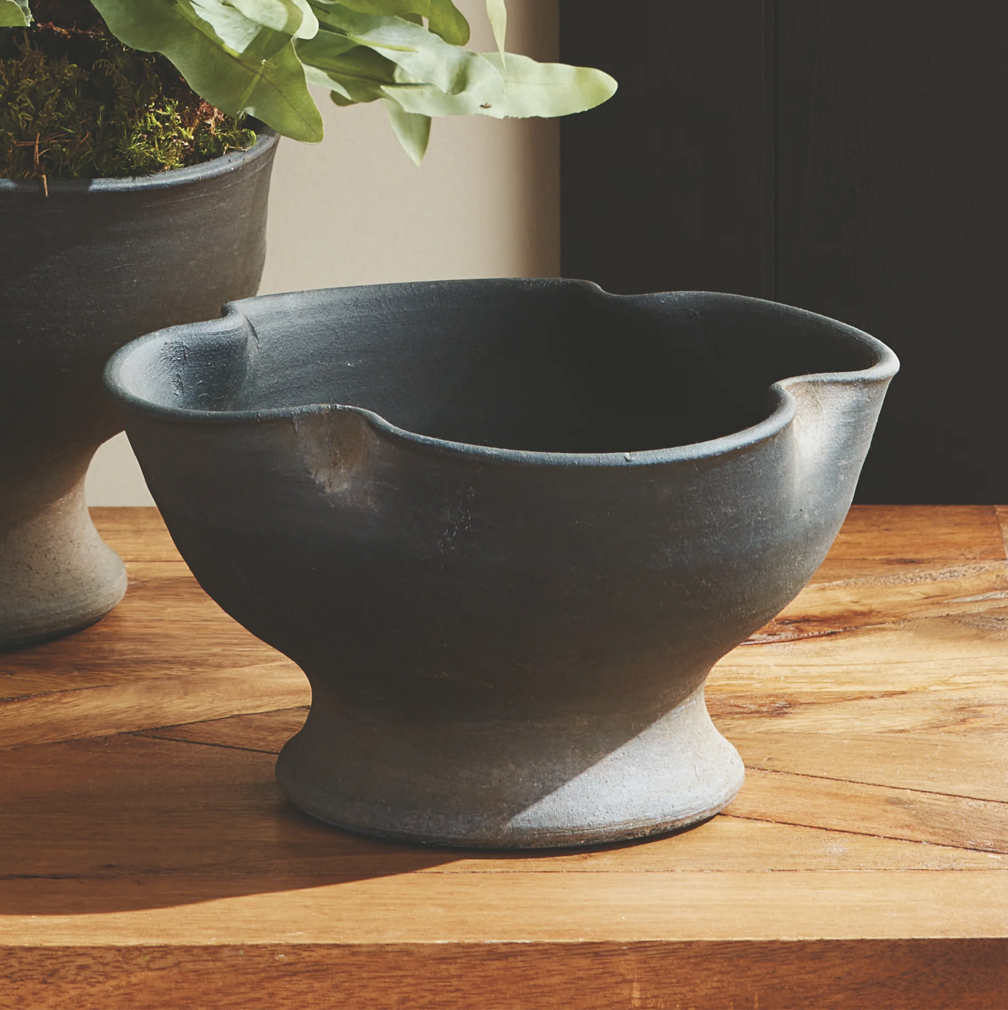 The Kaloa Bowl by Napa Home & Garden, a dark ceramic planter with a flared base and matte finish, rests on a wooden surface. In the background, part of another handcrafted planter with green foliage is visible.