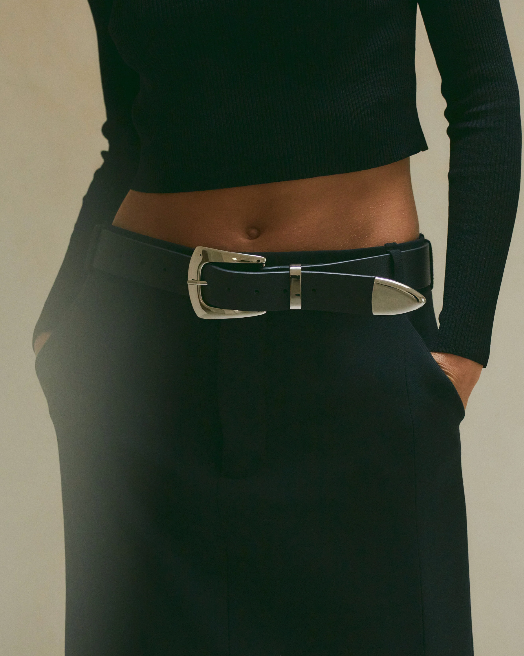 A person wearing a black ribbed long-sleeve top, black skirt or pants, and the B-Low The Belt Jordana Mini genuine leather Western belt with a large silver buckle stands with hands in their pockets.