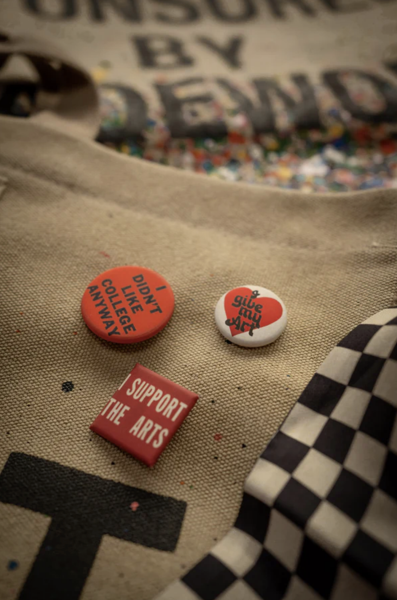 Three pins on the Made Worn Artist Tote: “DIDN’T LIKE COLLEGE ANYWAY,” “SUPPORT THE ARTS,” and a “SLICE MY WAY” graphic. The tan cotton canvas bag also features a black-and-white checkered pattern.