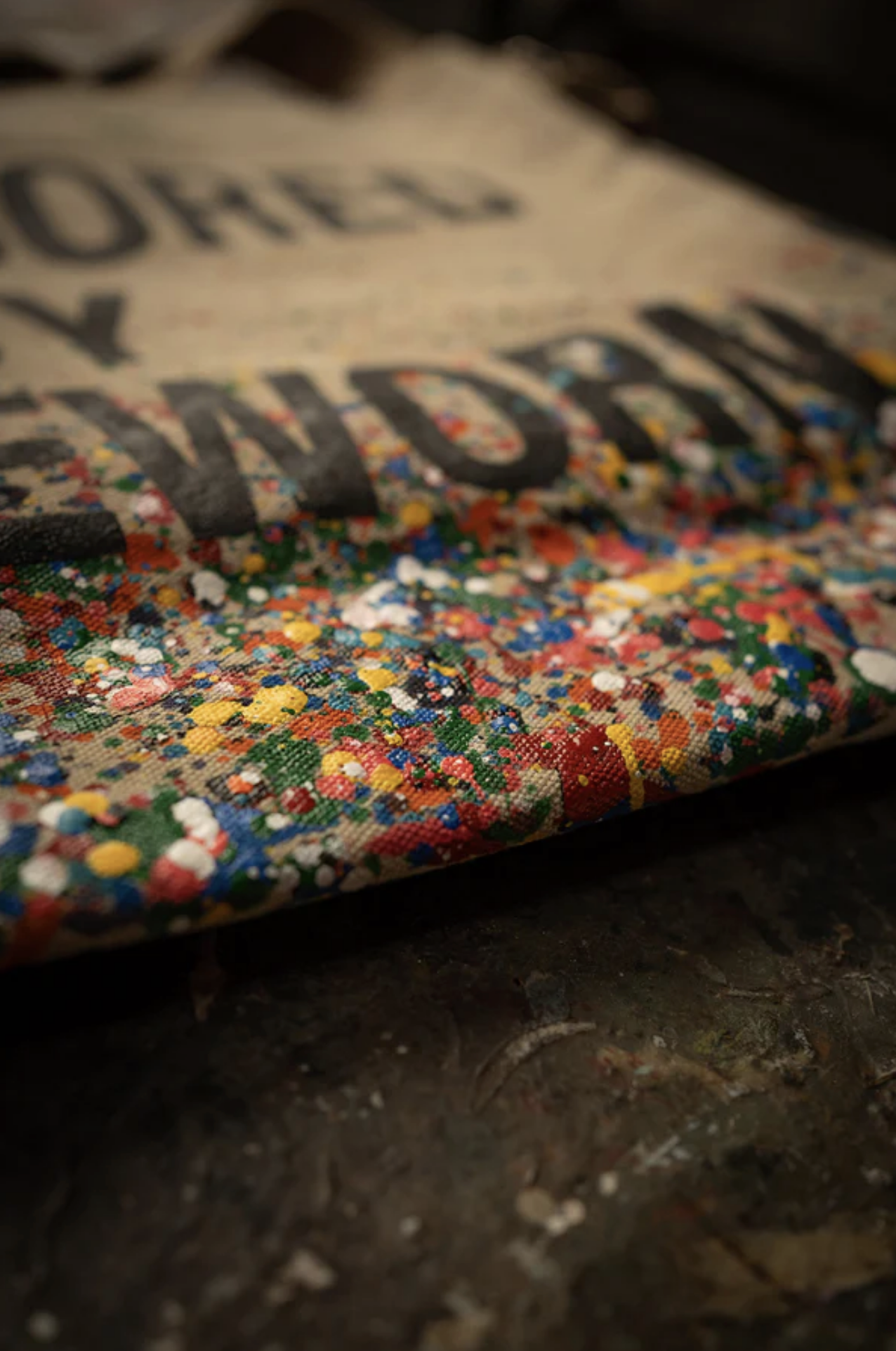 A close-up of the limited-edition Made Worn Artist Tote shows its vibrant paint-splattered cotton canvas, partially displaying bold black text on a tan background and highlighting the rich textured details.