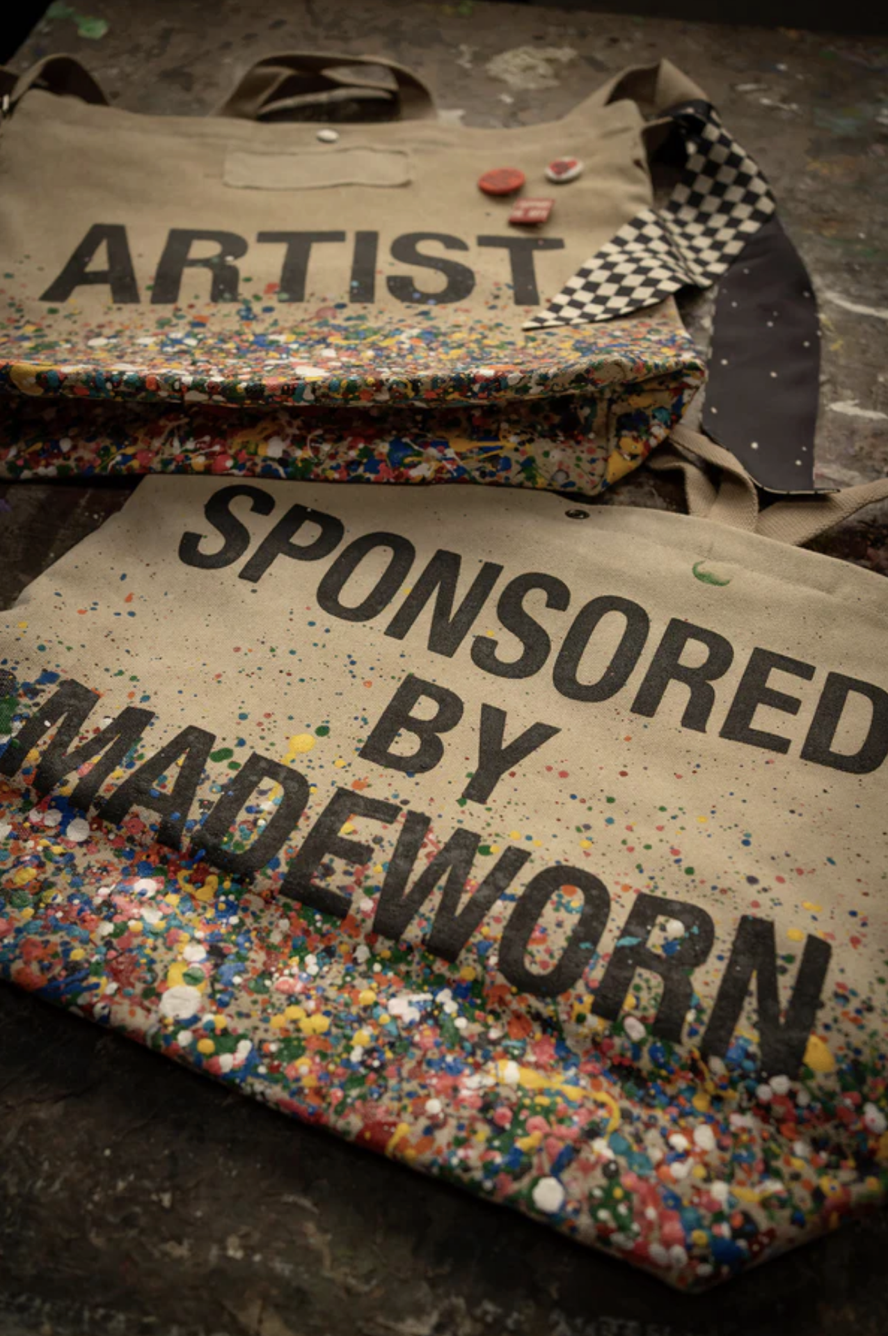 Two limited-edition beige cotton Artist Tote bags by Made Worn with colorful paint splatters—one features &quot;ARTIST,&quot; the other &quot;SPONSORED BY MADEWORN.&quot; The totes are displayed on a textured surface with custom buttons and a checkered cloth.