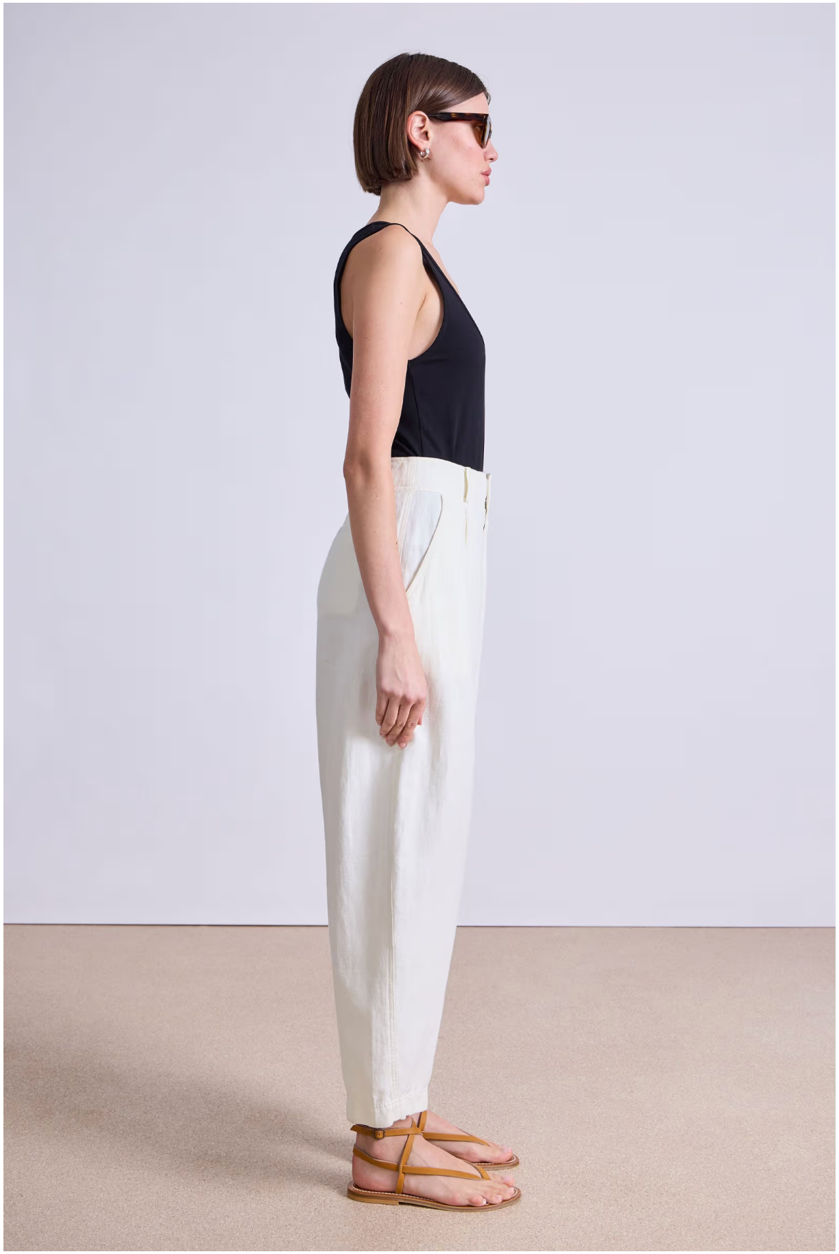 A person stands in profile against a plain background, wearing a black sleeveless top, Apiece Apart Bari Crop Trouser, brown sandals, and sunglasses—a minimalist and modern look.