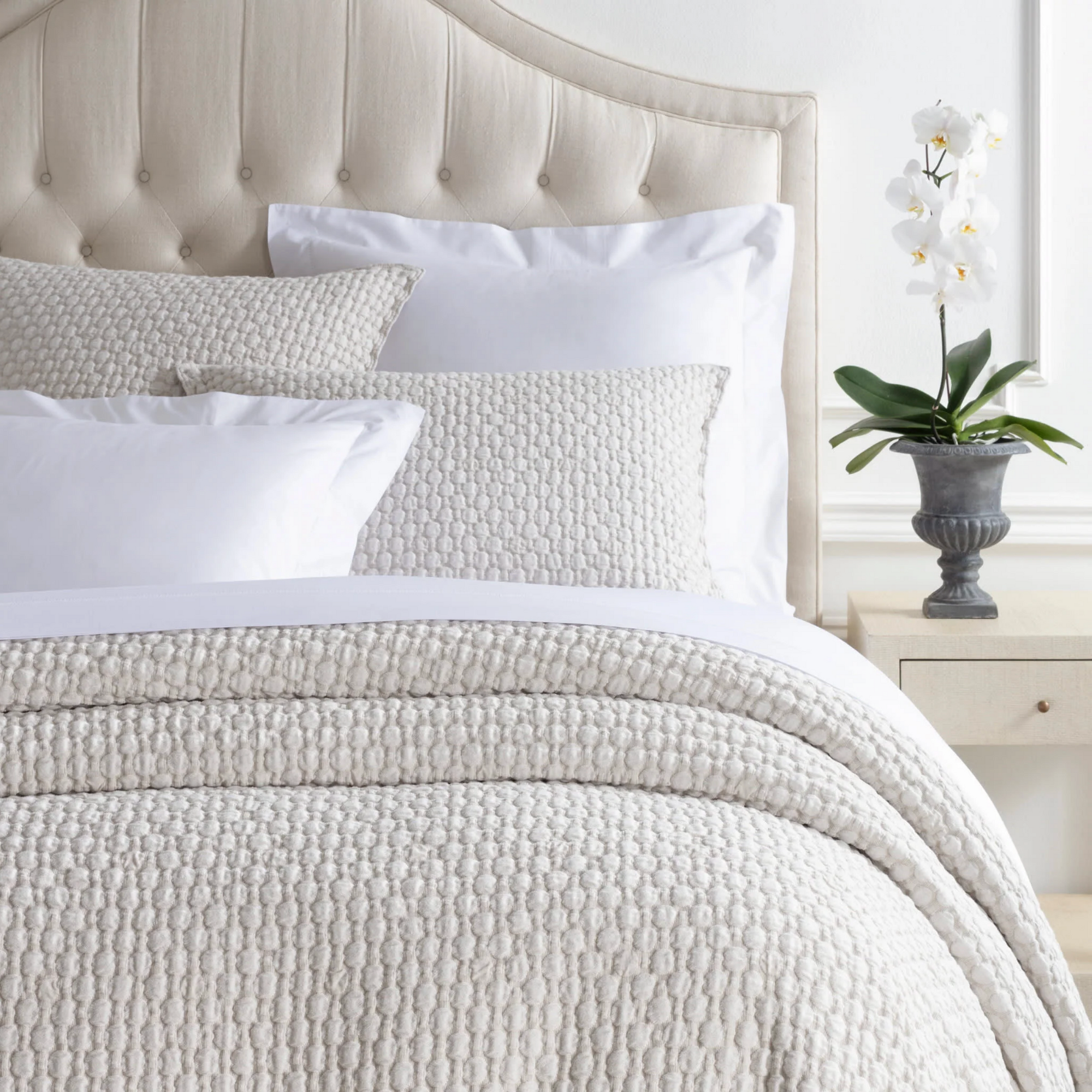 A neatly made bed features a beige Pinecone Hill Lodi Matelasse Coverlet, white pillows, and a tufted beige headboard. A potted white orchid rests on a light nightstand beside the bed against a white wall.