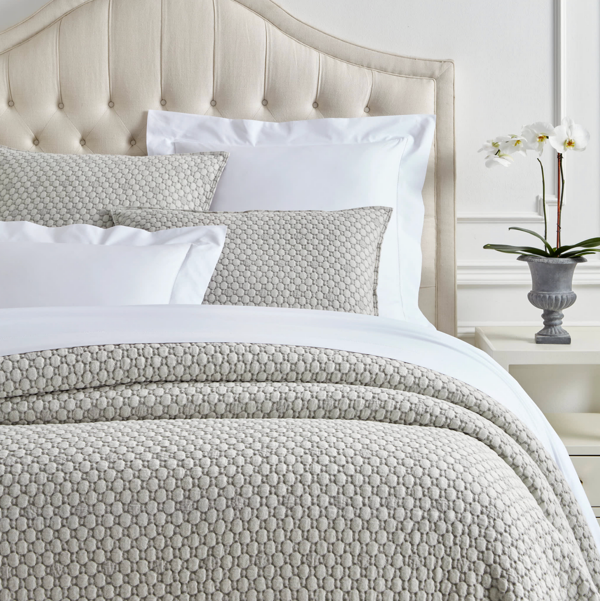 A neatly made bed features the Pinecone Hill Lodi Matelasse Coverlet and pillows in light gray, a tufted beige headboard, a white nightstand with a gray pot and white orchid, set against white paneled walls.