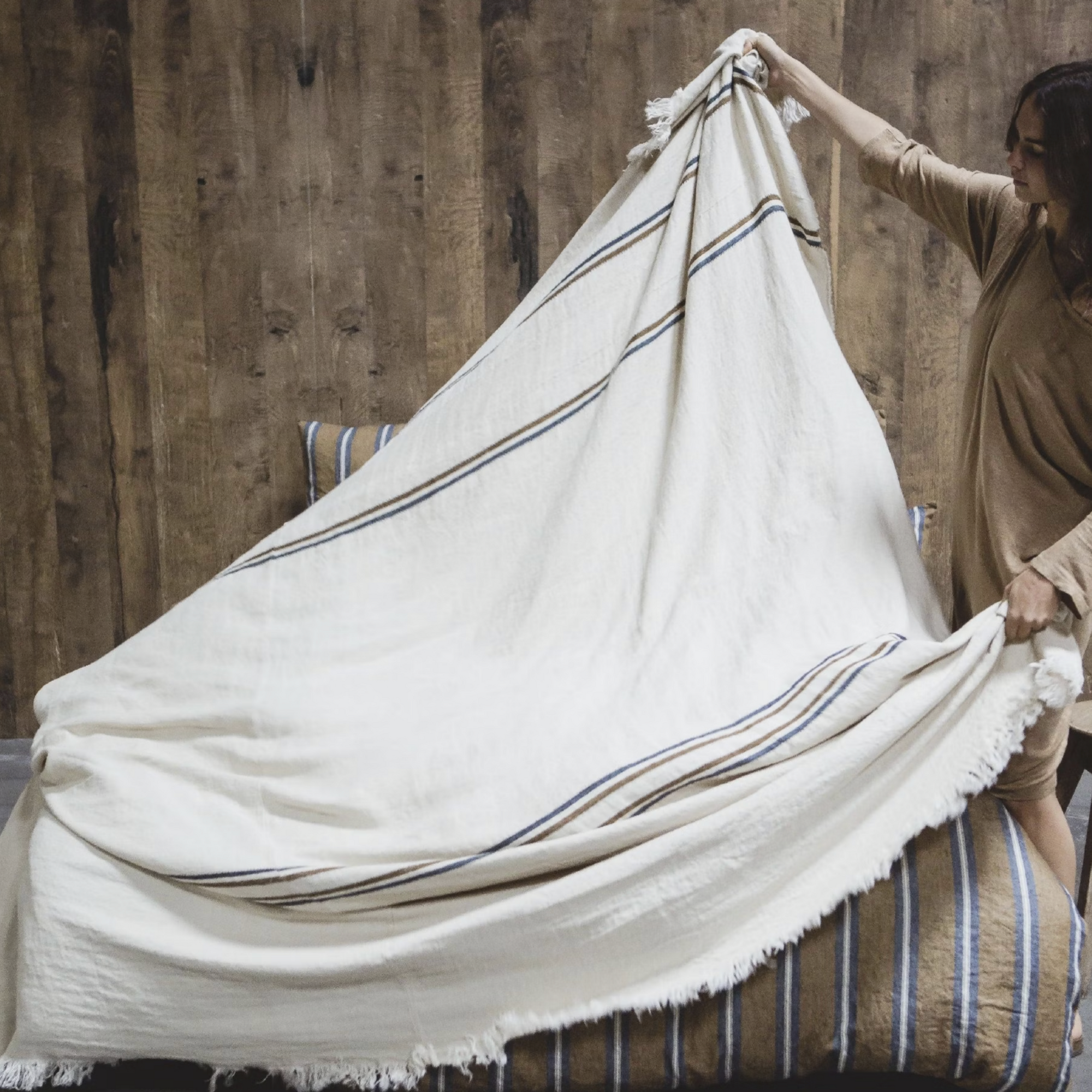 A person spreads the Libeco Home Auburn Coverlet, a large cream-colored towel with navy and bronze stripes, over a bed with striped bedding, set against a wooden wall background.