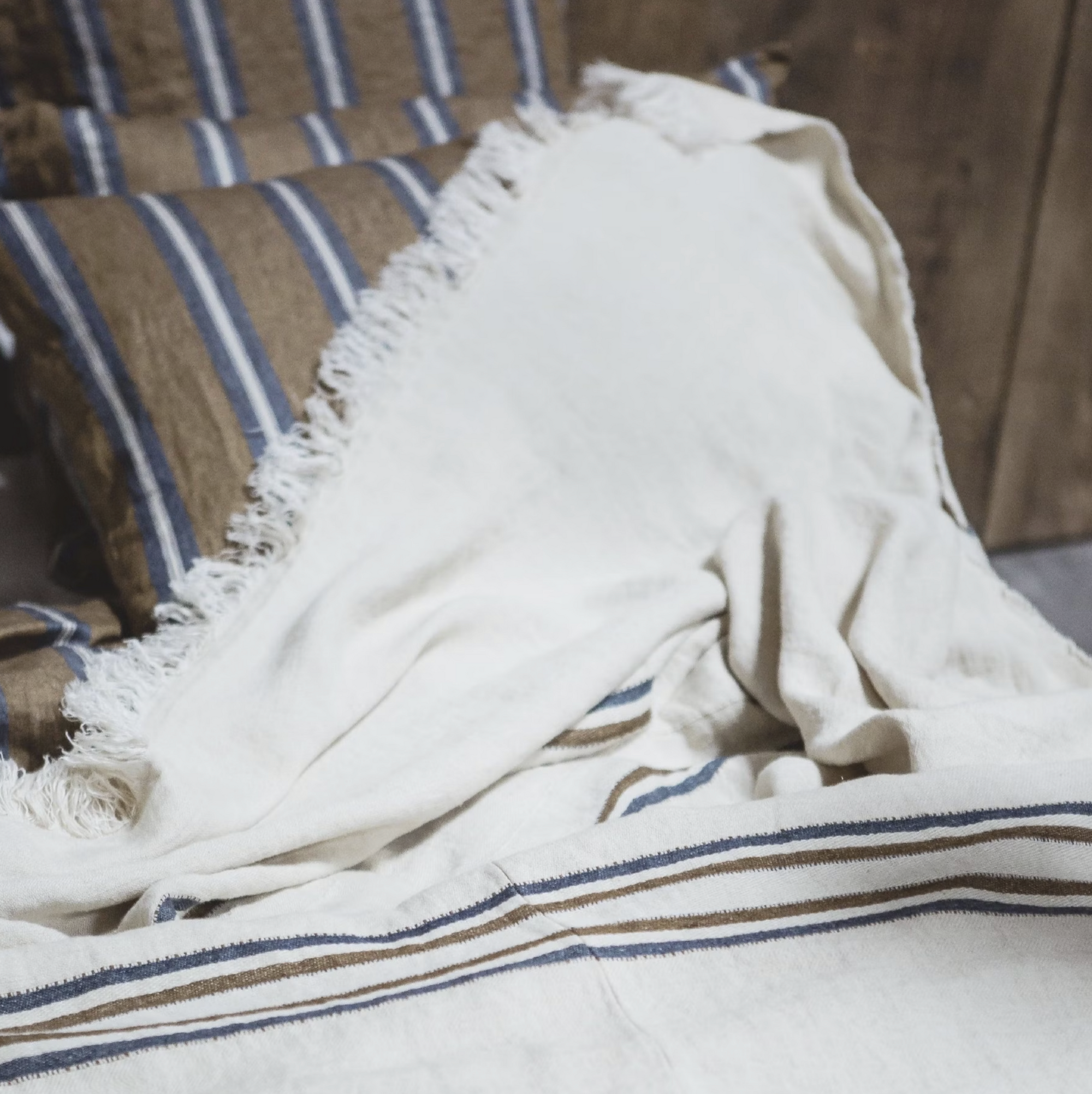 The Libeco Home Auburn Coverlet, in cream with navy and bronze stripes, is draped over a bed, coordinating with striped pillows and complementing the wooden wall behind them.
