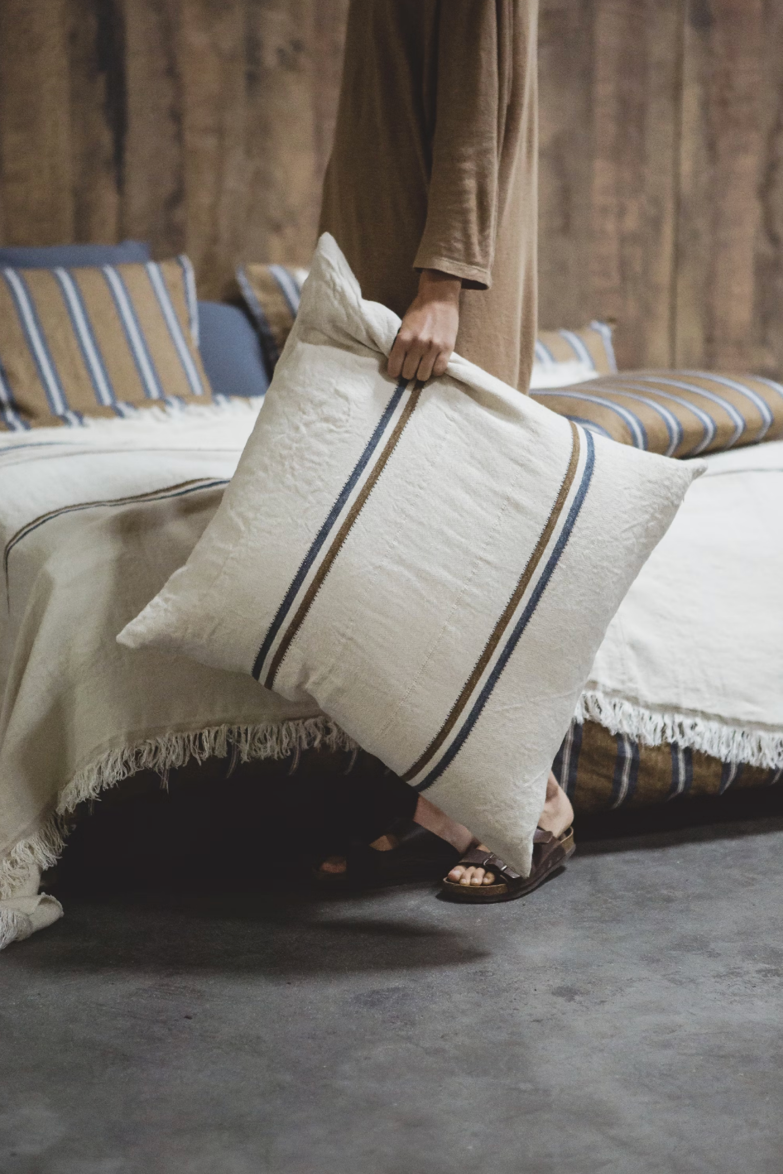 Wearing a brown outfit and sandals, a person holds the Libeco Home Auburn Pillow—white with navy and bronze stripes—beside a bed adorned with matching striped, fringed bedding in a rustic room.