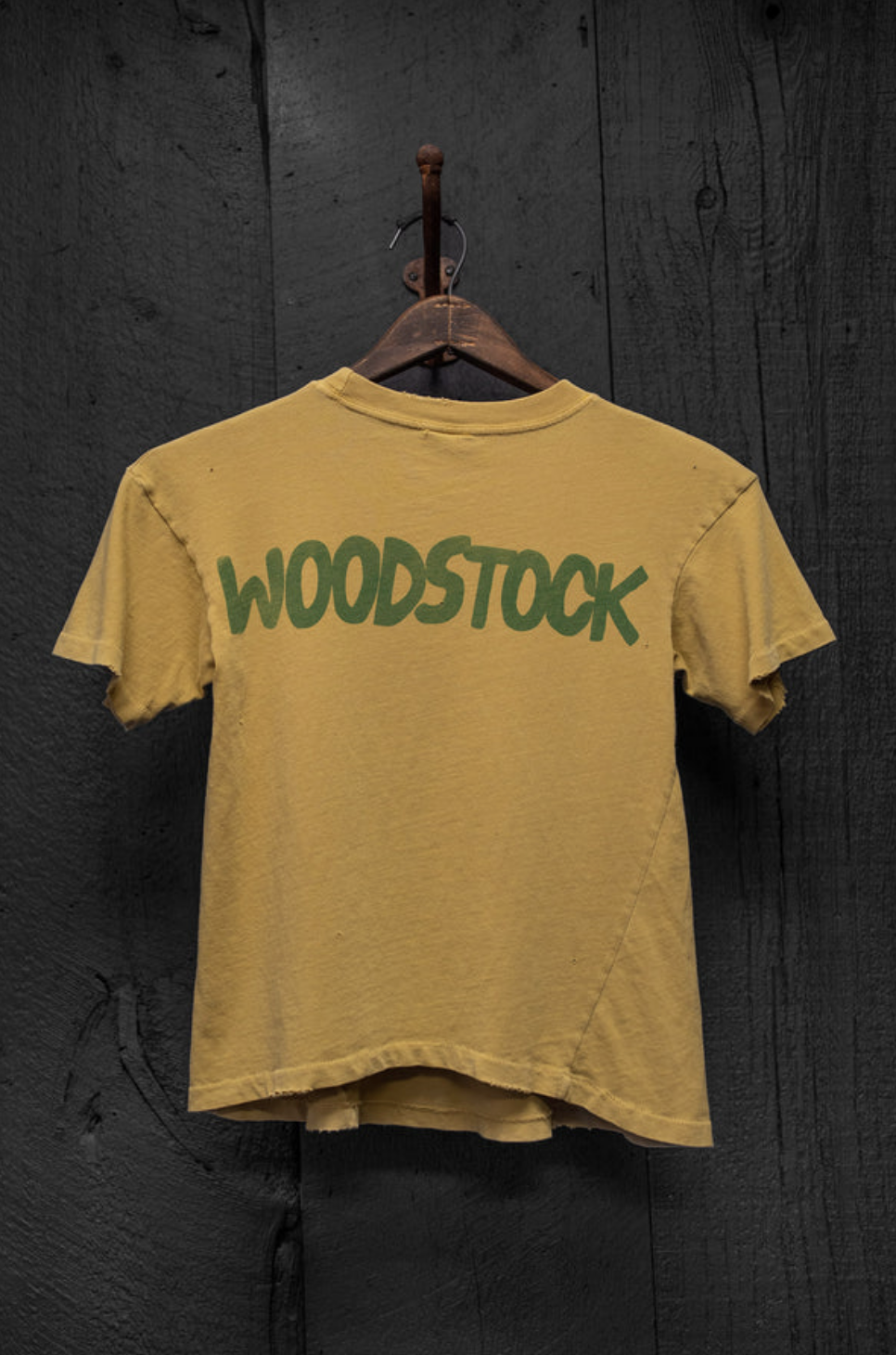 The Made Worn Woodstock Crop Tee Mustard is a yellow vintage-fit top with "WOODSTOCK" in green block letters on the back, displayed on a wooden hanger against a dark wooden background.